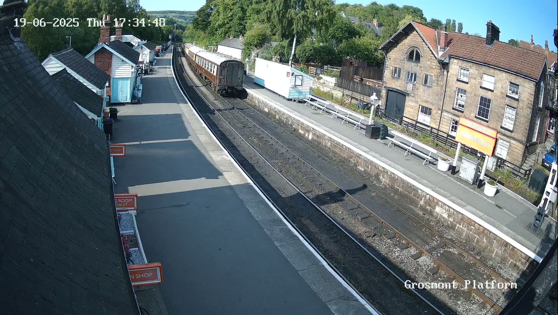 A train departs a railway station on a sunny day, passing alongside small buildings and a larger stone structure.