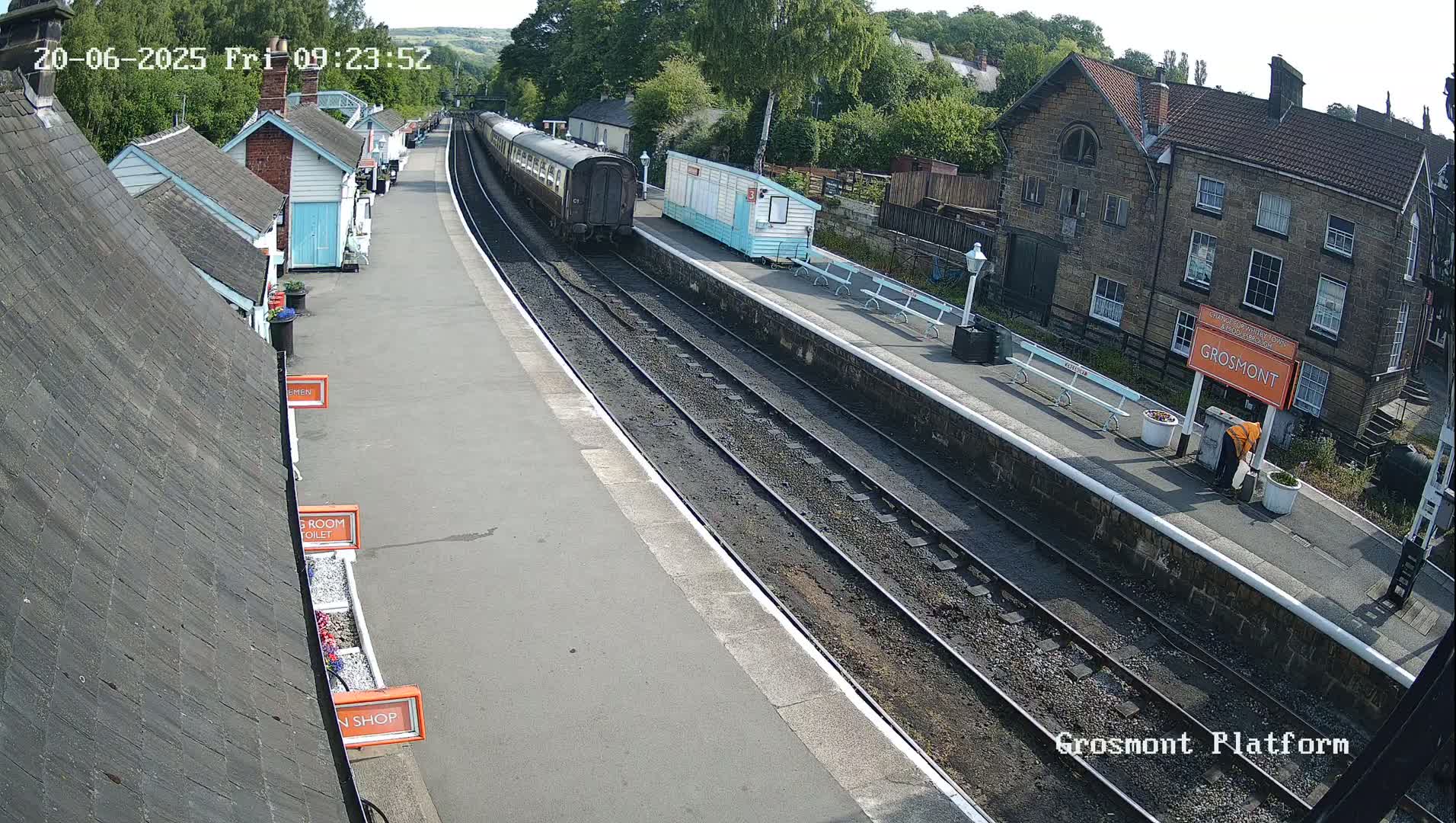 A train sits at a railway station platform on a sunny day, with several small buildings lining the platform and a larger stone building visible in the background.