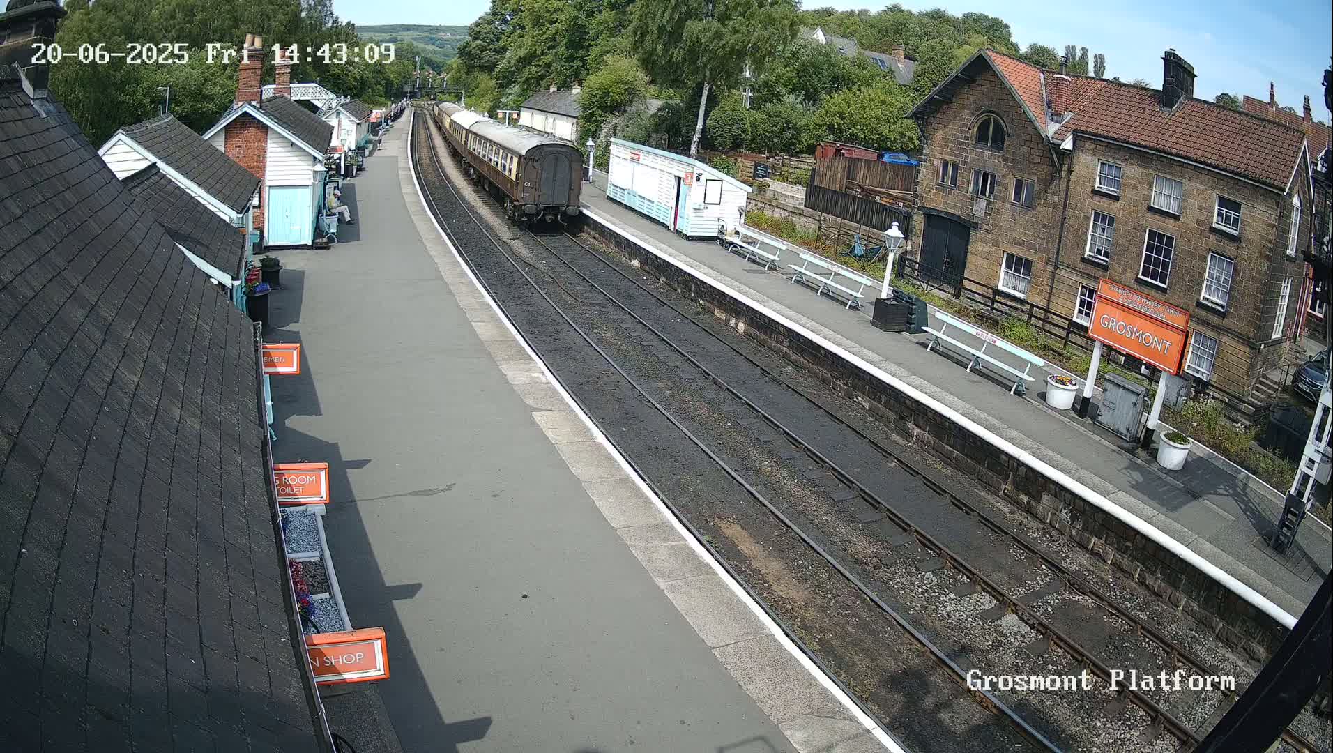 A train is arriving at a railway station on a sunny day, with several buildings visible alongside the platform.