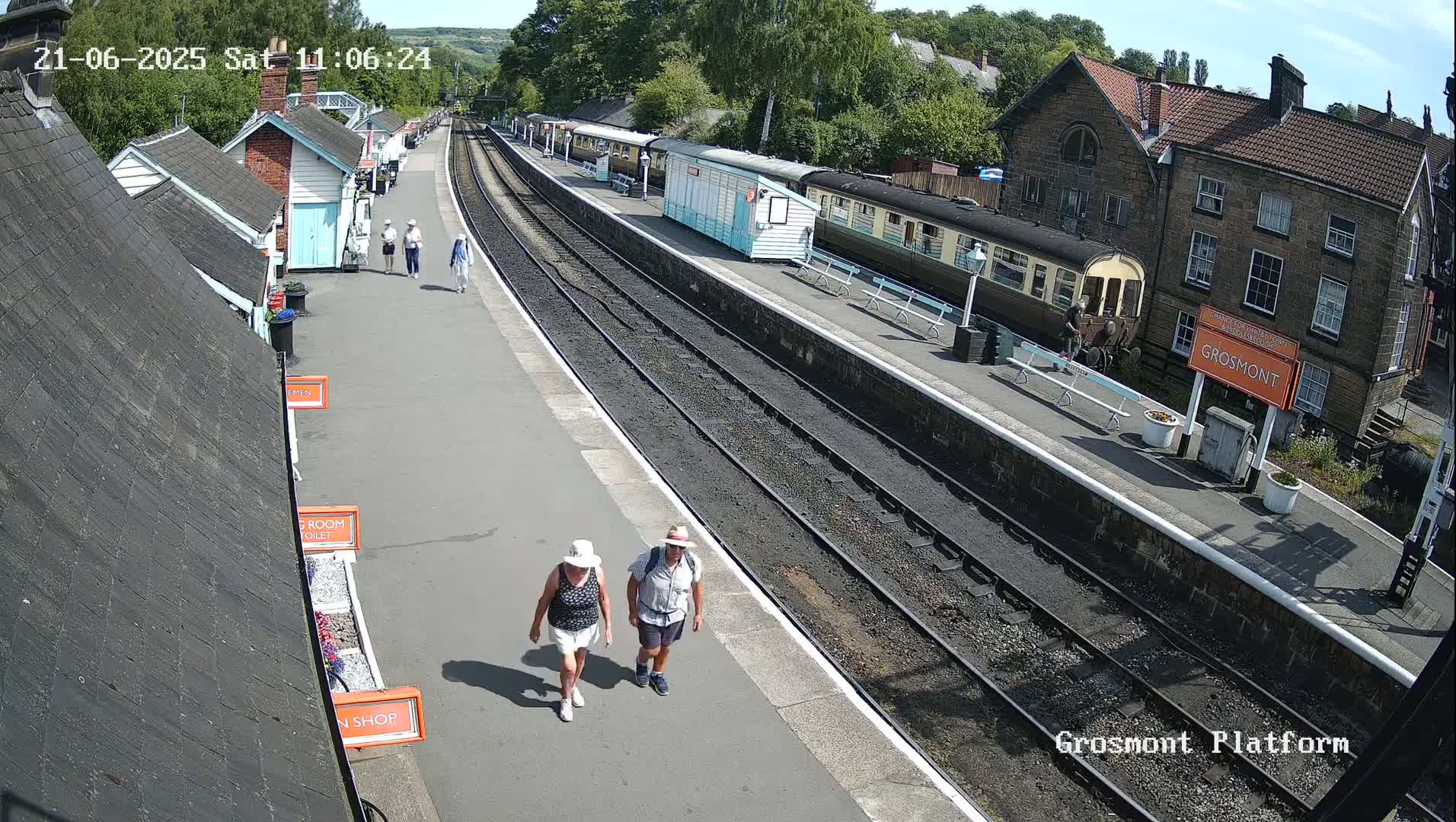 A sunny day at a train station shows several people walking on a platform next to a train and several small buildings.