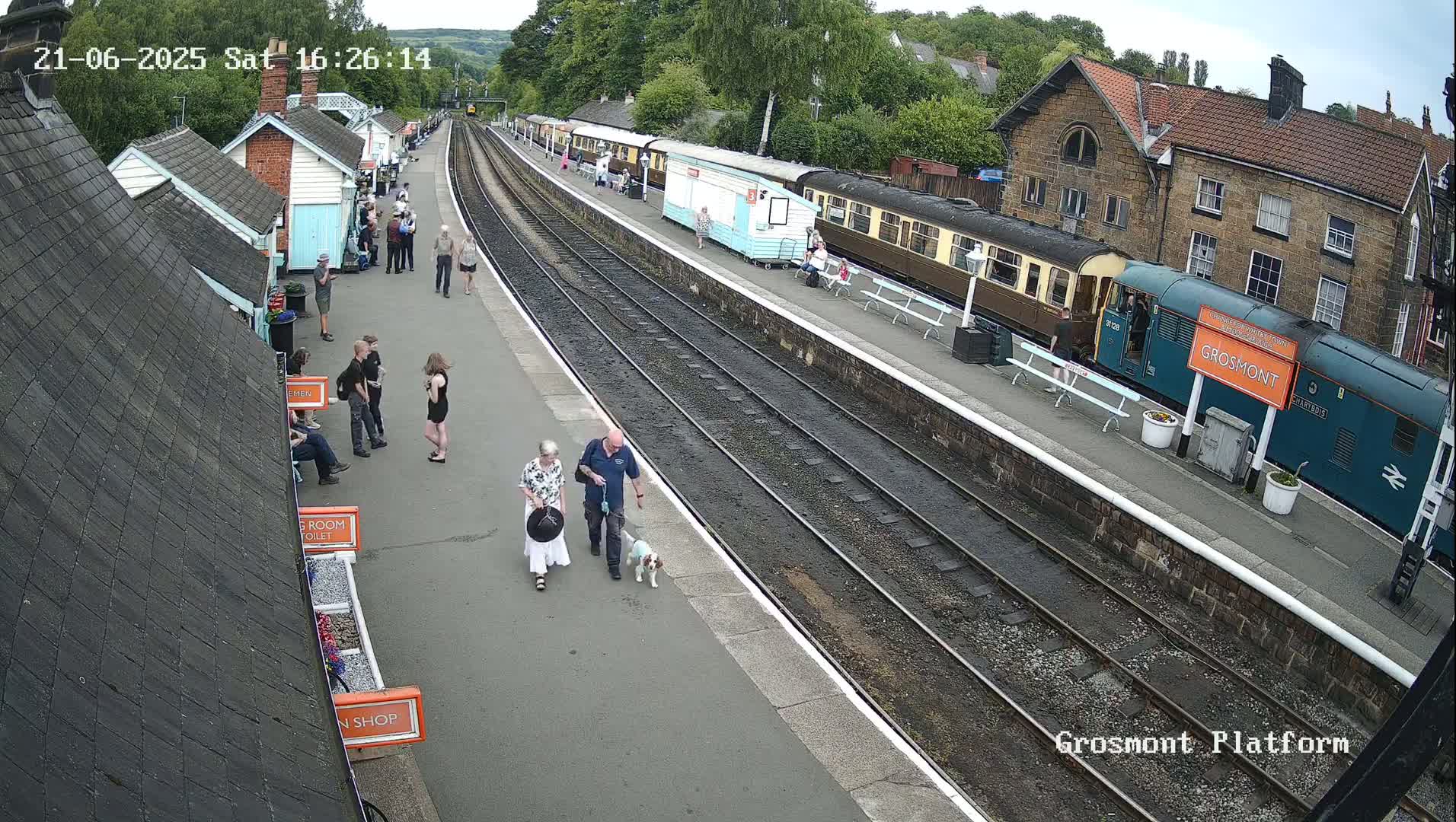 A train station platform with several people, some walking a dog, near a train and several small buildings under a partly cloudy sky.