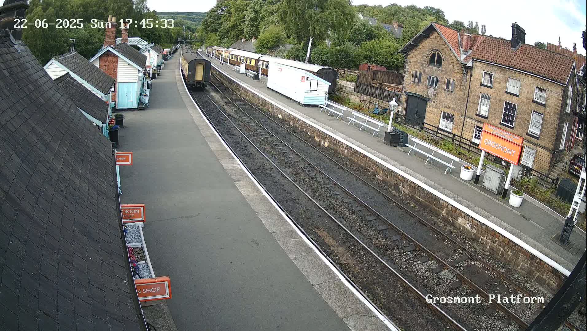 A train sits at a station platform on a partly sunny day, with several passenger cars parked nearby and small buildings lining the platform.