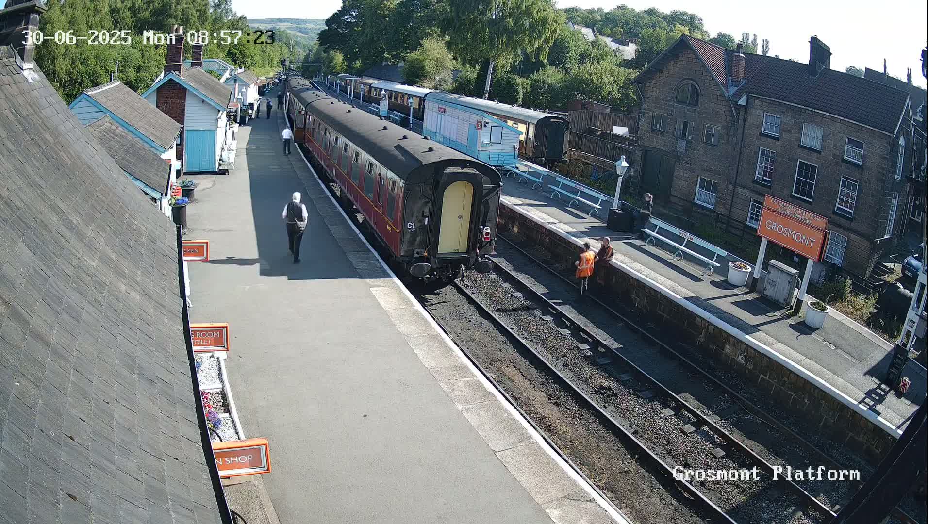 A train is at a station platform on a sunny day, with several people walking nearby and a few standing near the train.