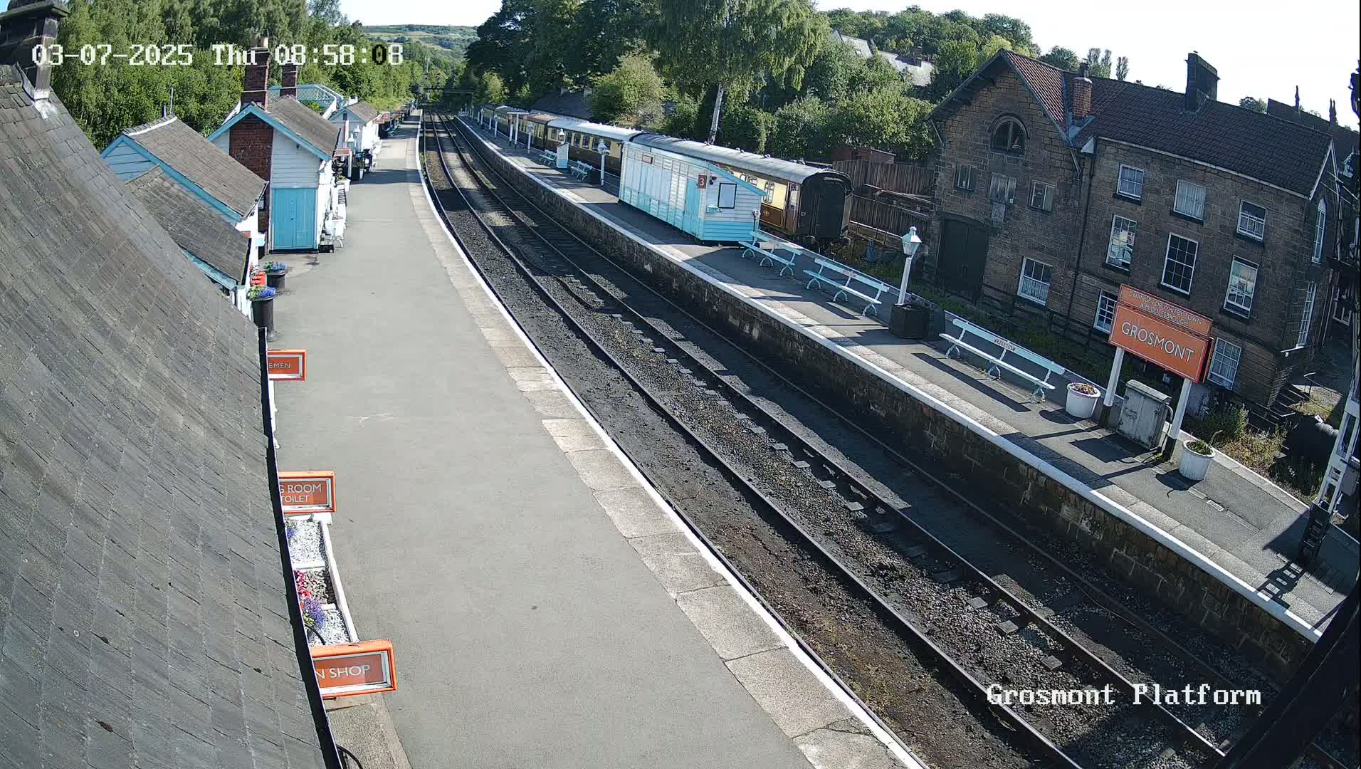 A sunny day at a train station shows a railway platform with several train cars, small buildings, and a larger stone building in the background.