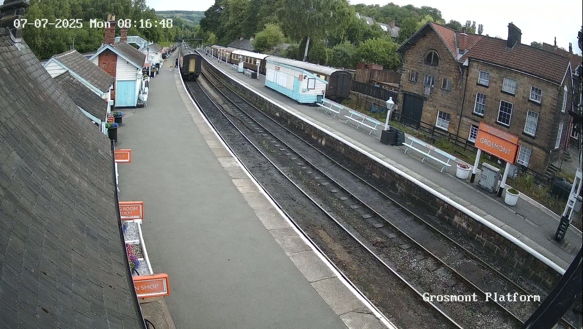 A train sits at a station platform on a partly sunny day, with several small buildings along the platform and a larger stone building visible beyond the tracks.