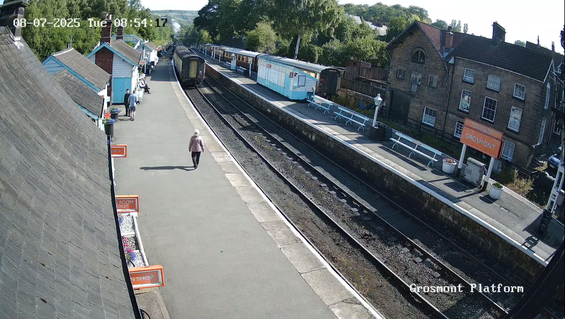 A train sits at a station platform on a sunny day, with a few people walking on the platform.