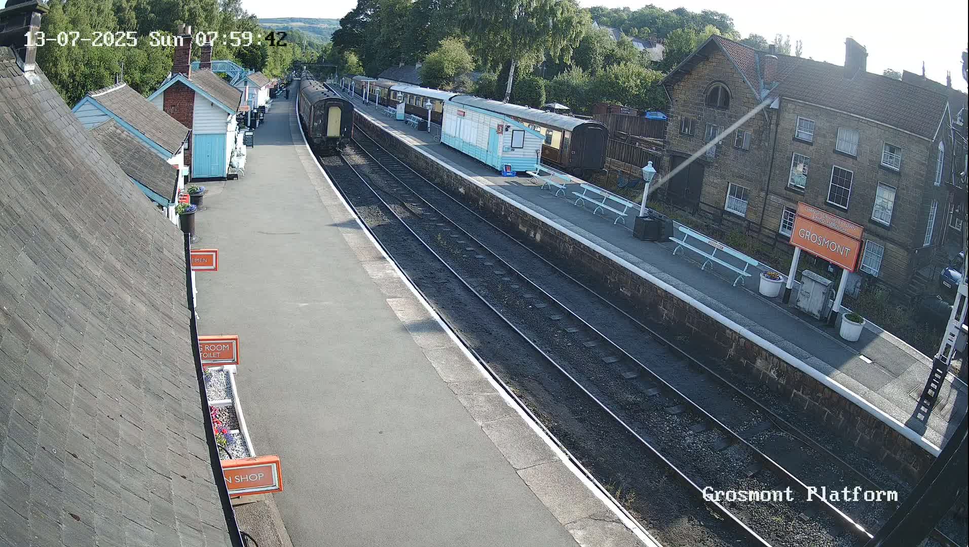 A train sits at a station platform on a sunny day, with several small buildings and a larger stone structure visible nearby.