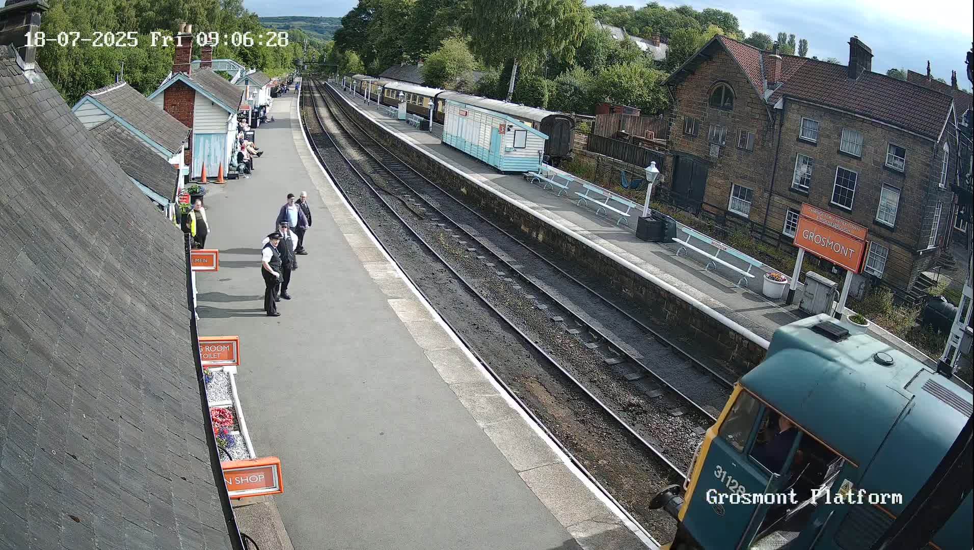 A train arrives at a railway station on a sunny day, with several people standing on the platform near some buildings.