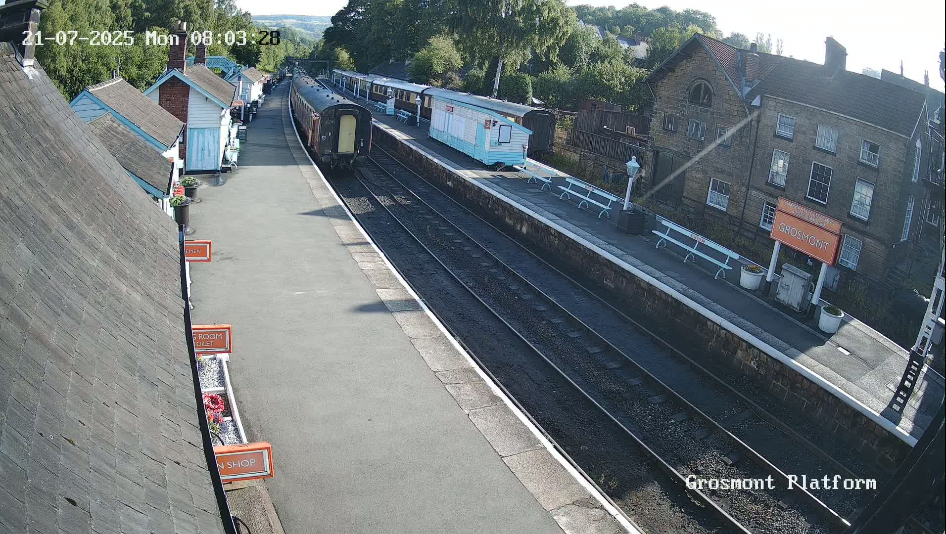 A train sits at a station platform on a sunny day, with several small buildings and a larger stone building visible in the background.