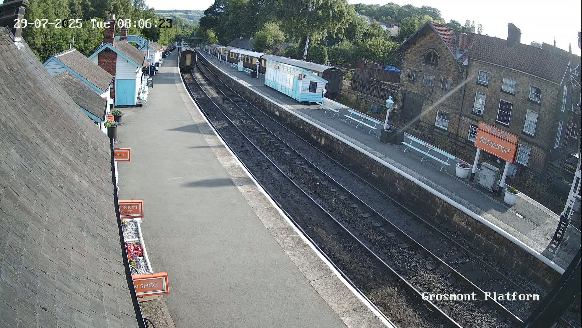 A train sits at a station platform on a sunny day, with several small buildings visible alongside the tracks.