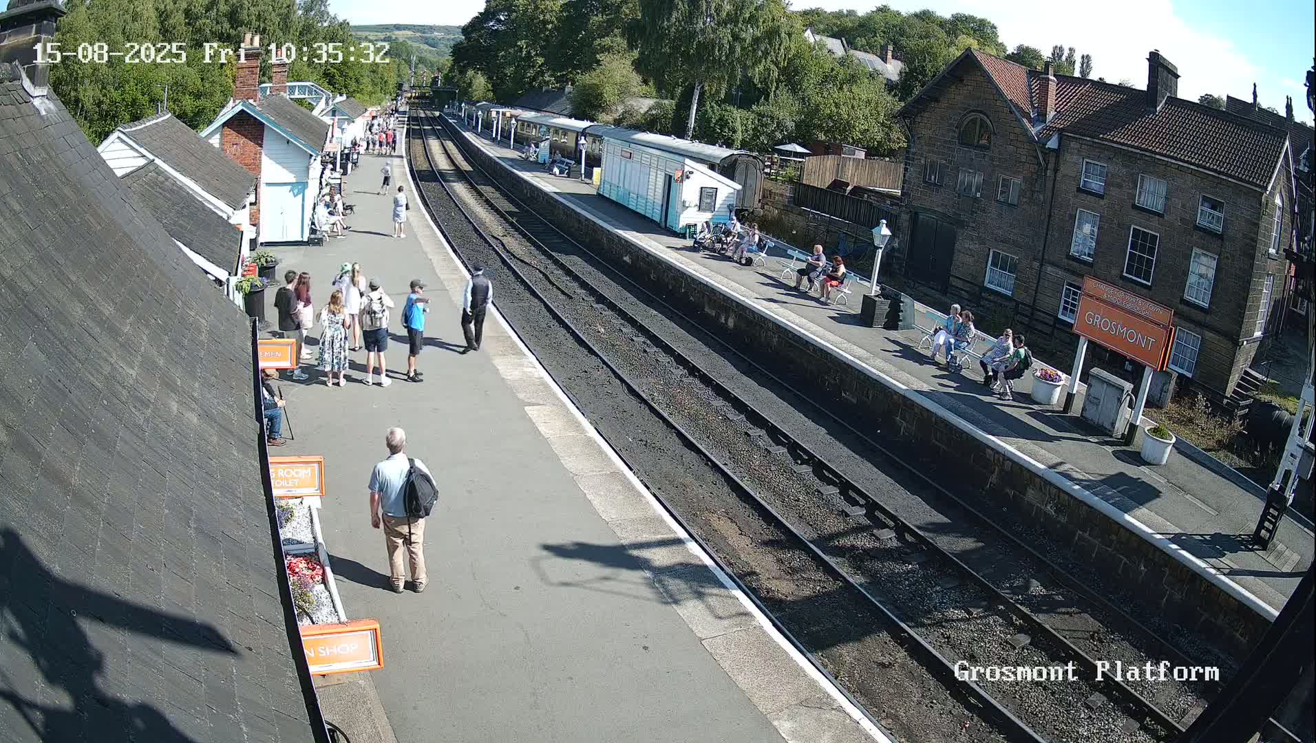 A train sits at a station platform on a sunny day, with several small buildings visible alongside the tracks.