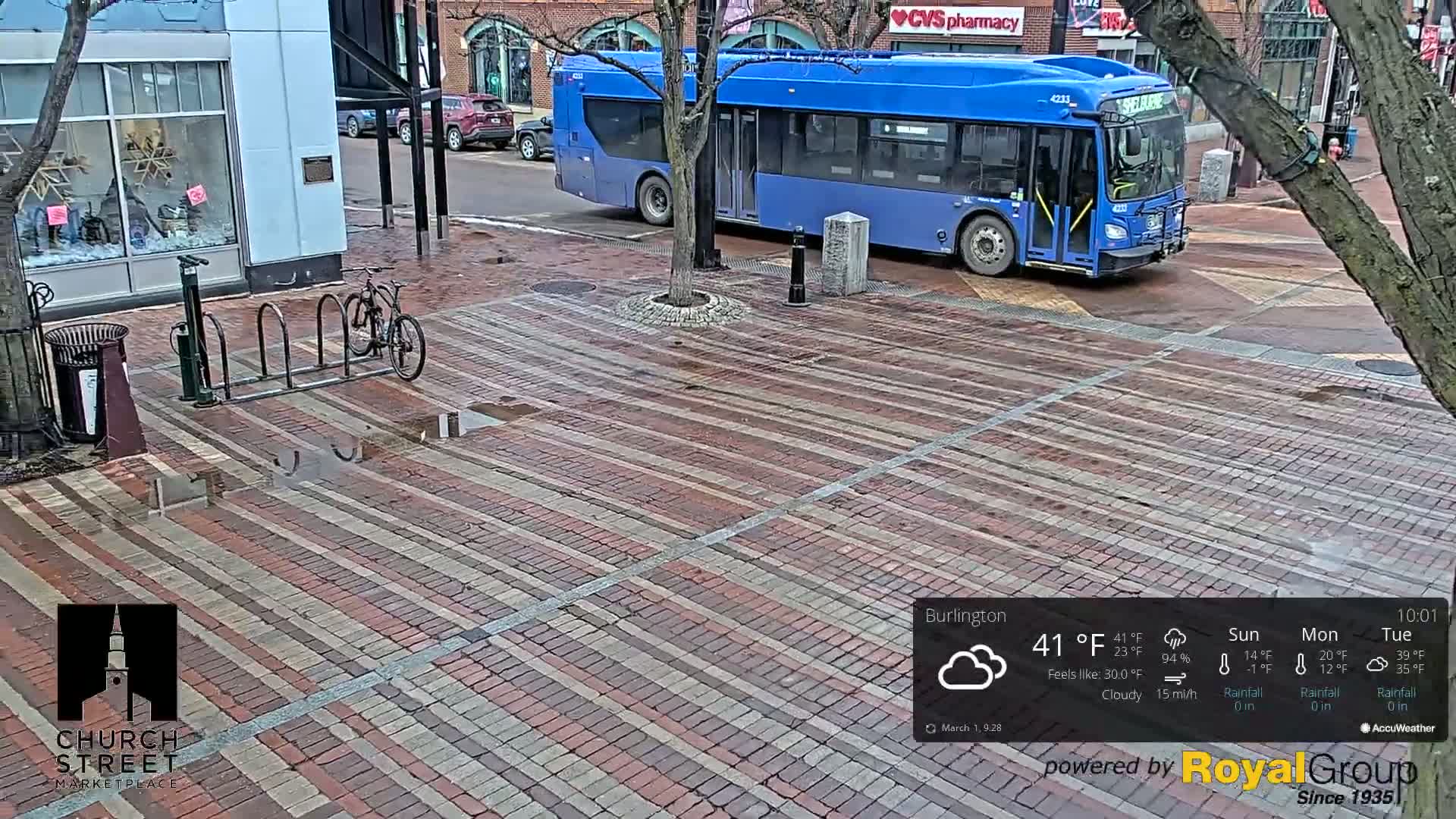 A brick-paved city street with a blue bus, a bicycle rack, and puddles, under cloudy skies.