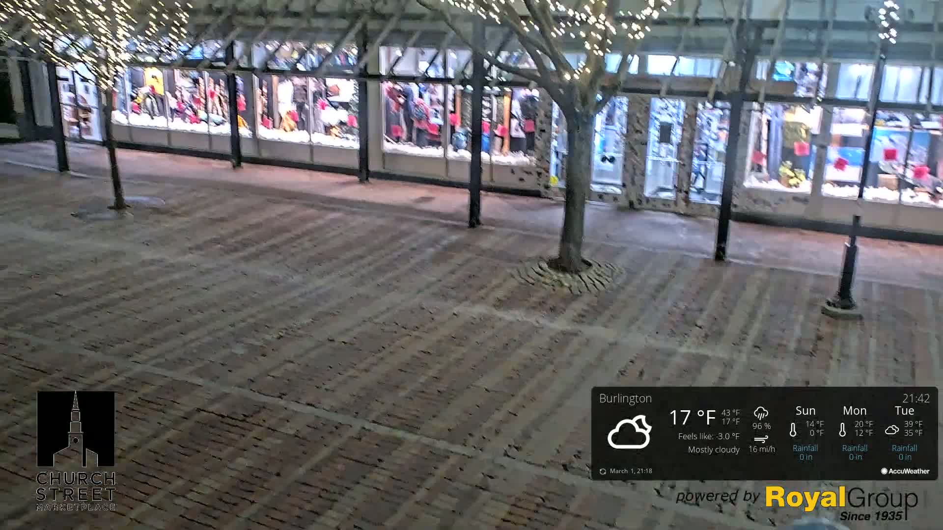 A mostly cloudy night scene shows a brick paved area in front of shops with holiday lights strung above.