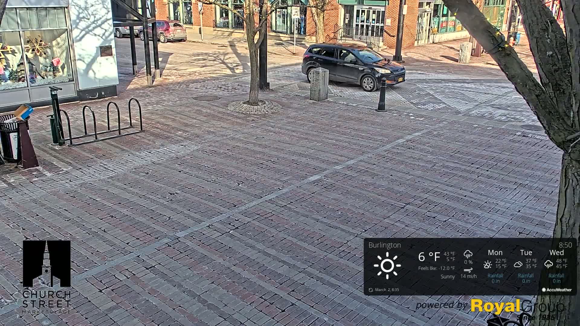 A brick paved area with a parked dark-colored SUV, a bike rack, and a tree, under sunny conditions.