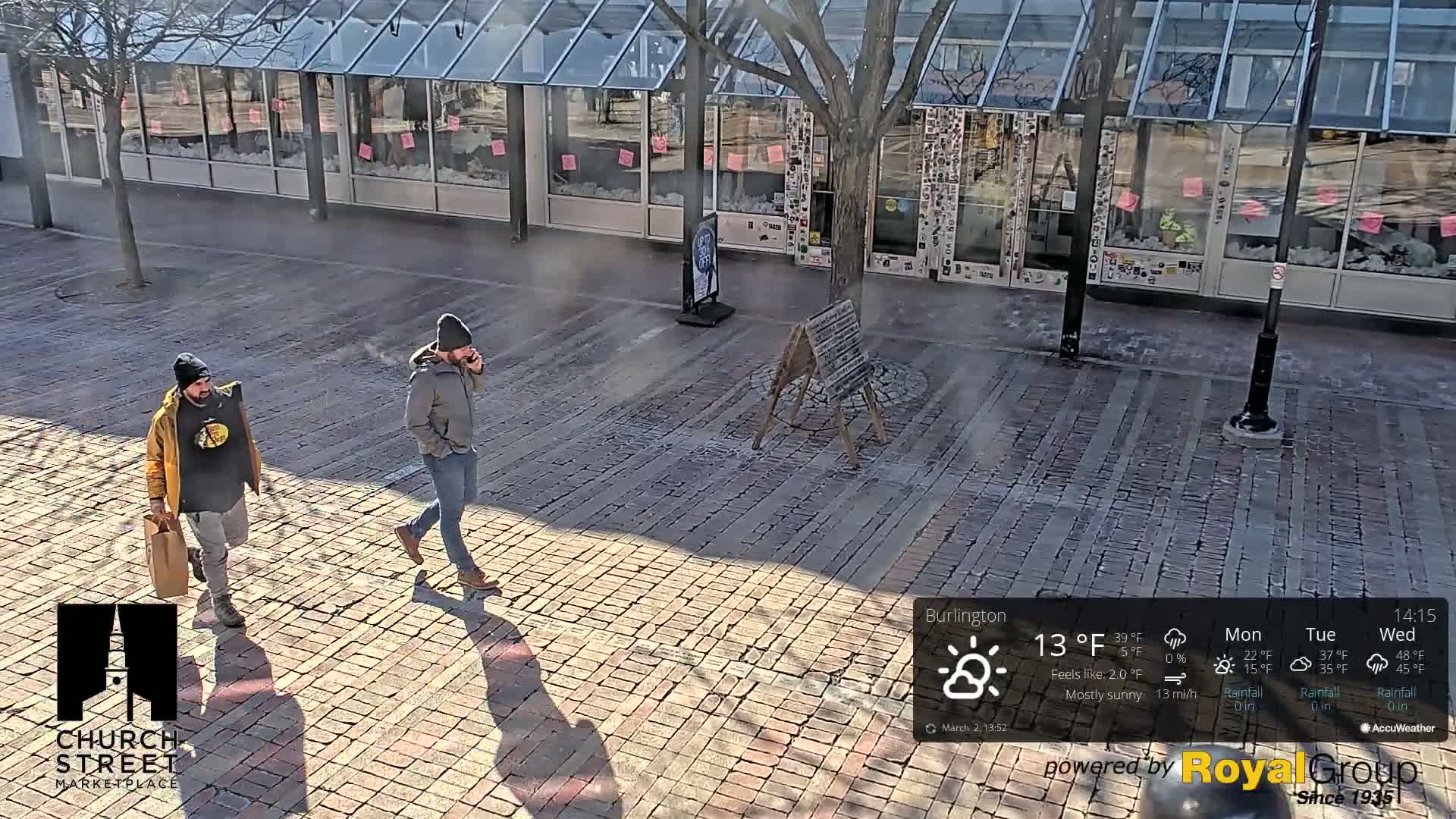 Two men walk on a brick-paved street in mostly sunny weather, past a building with large windows.