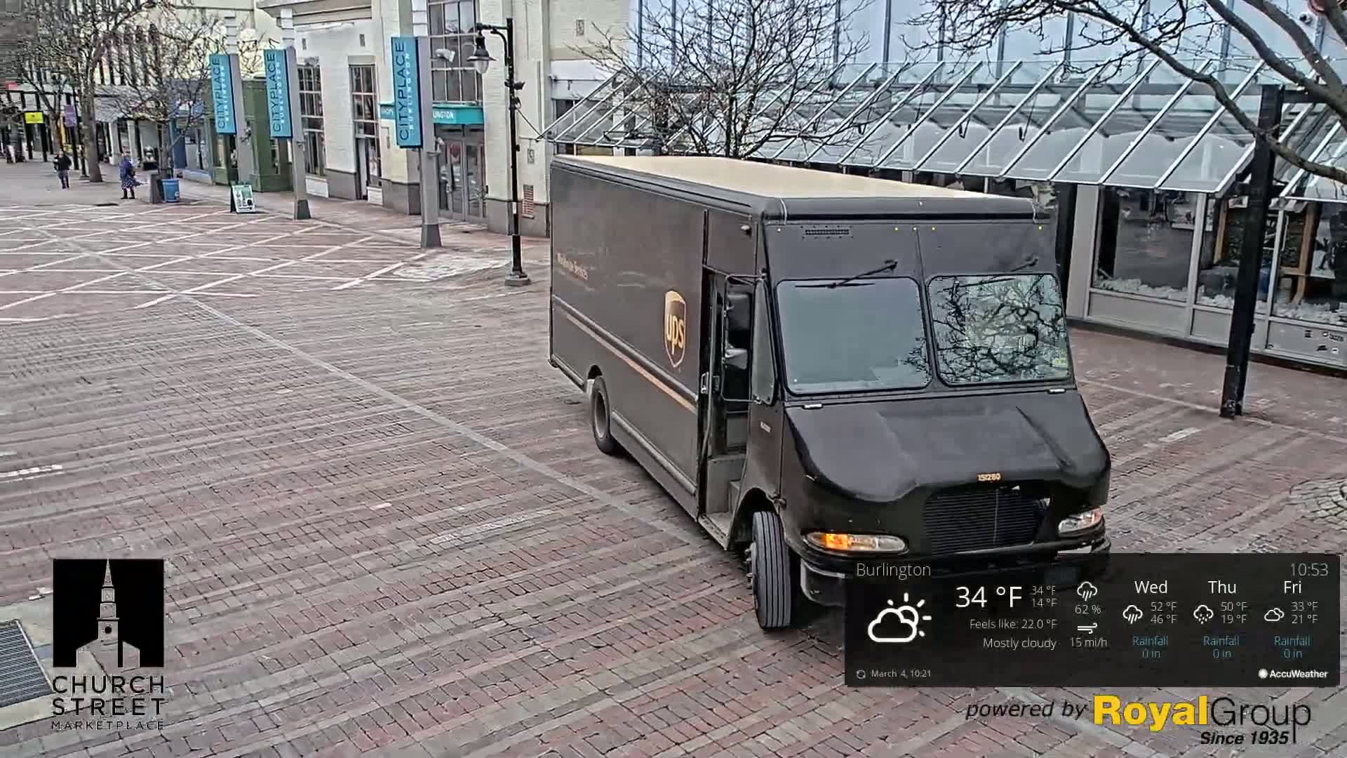 A brown UPS delivery truck is parked on a brick-paved street in mostly cloudy weather with a 15 mph wind.