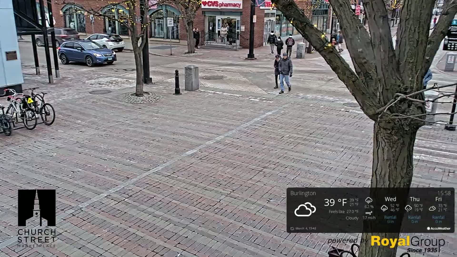 A brick paved street with a few pedestrians and parked cars, under cloudy skies.