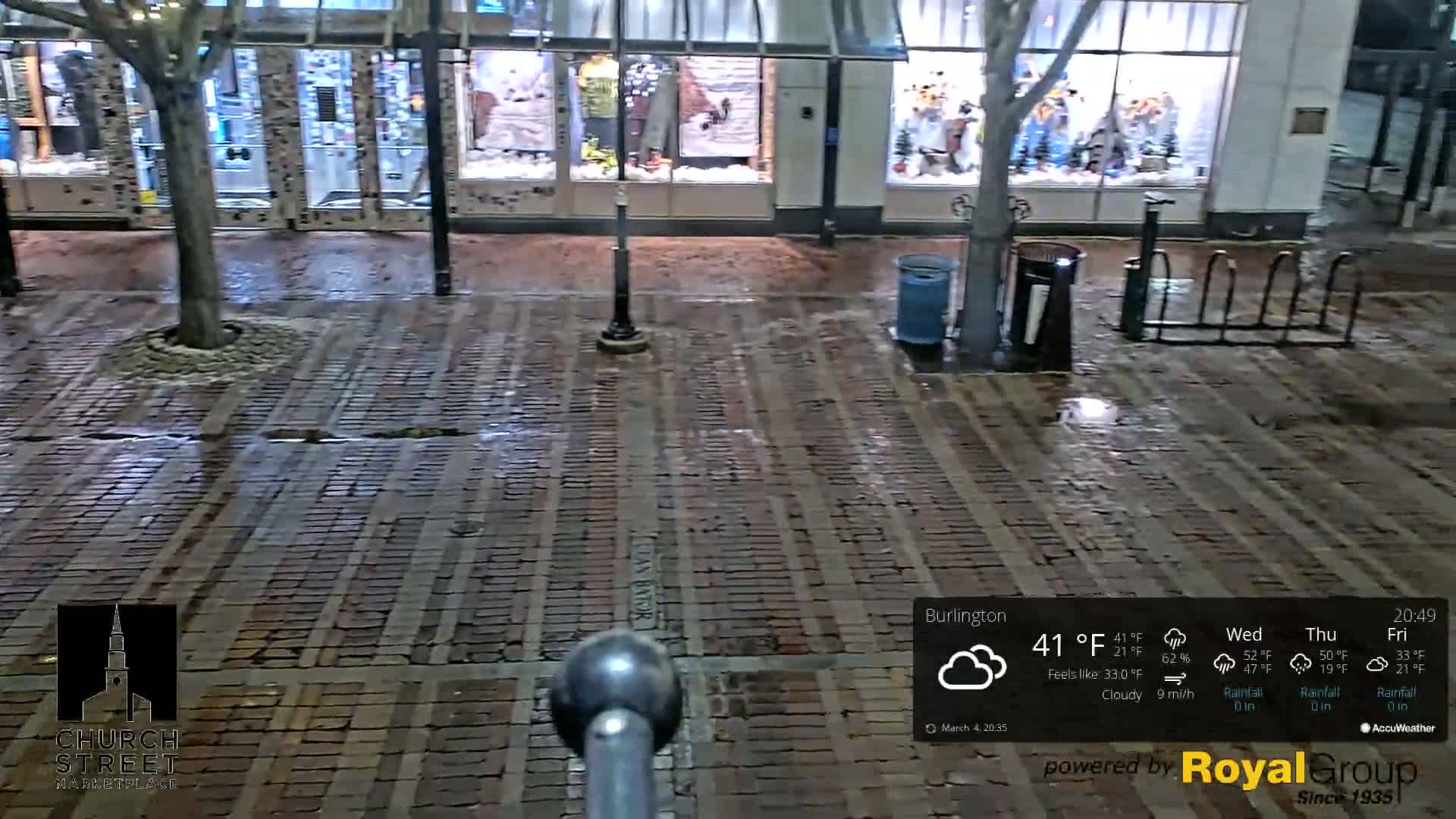 A wet brick street at night, under cloudy skies, is seen in front of storefronts.