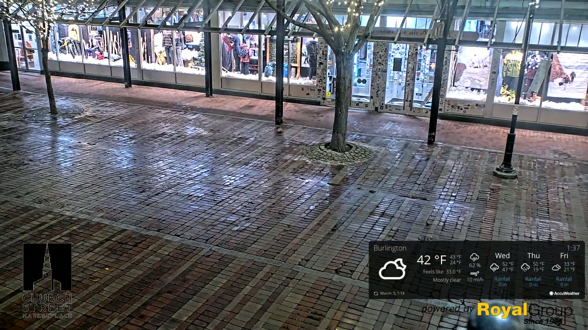 A mostly clear night shows a brick-paved walkway in front of storefronts with holiday decorations, a tree in the center, and some light snow on the ground.