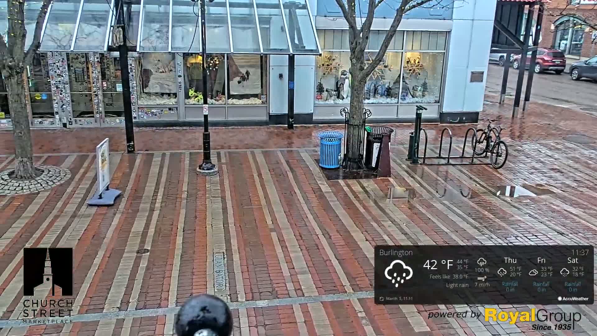A wet brick street with a bicycle rack and a few trash cans in front of storefronts, under a light rain.