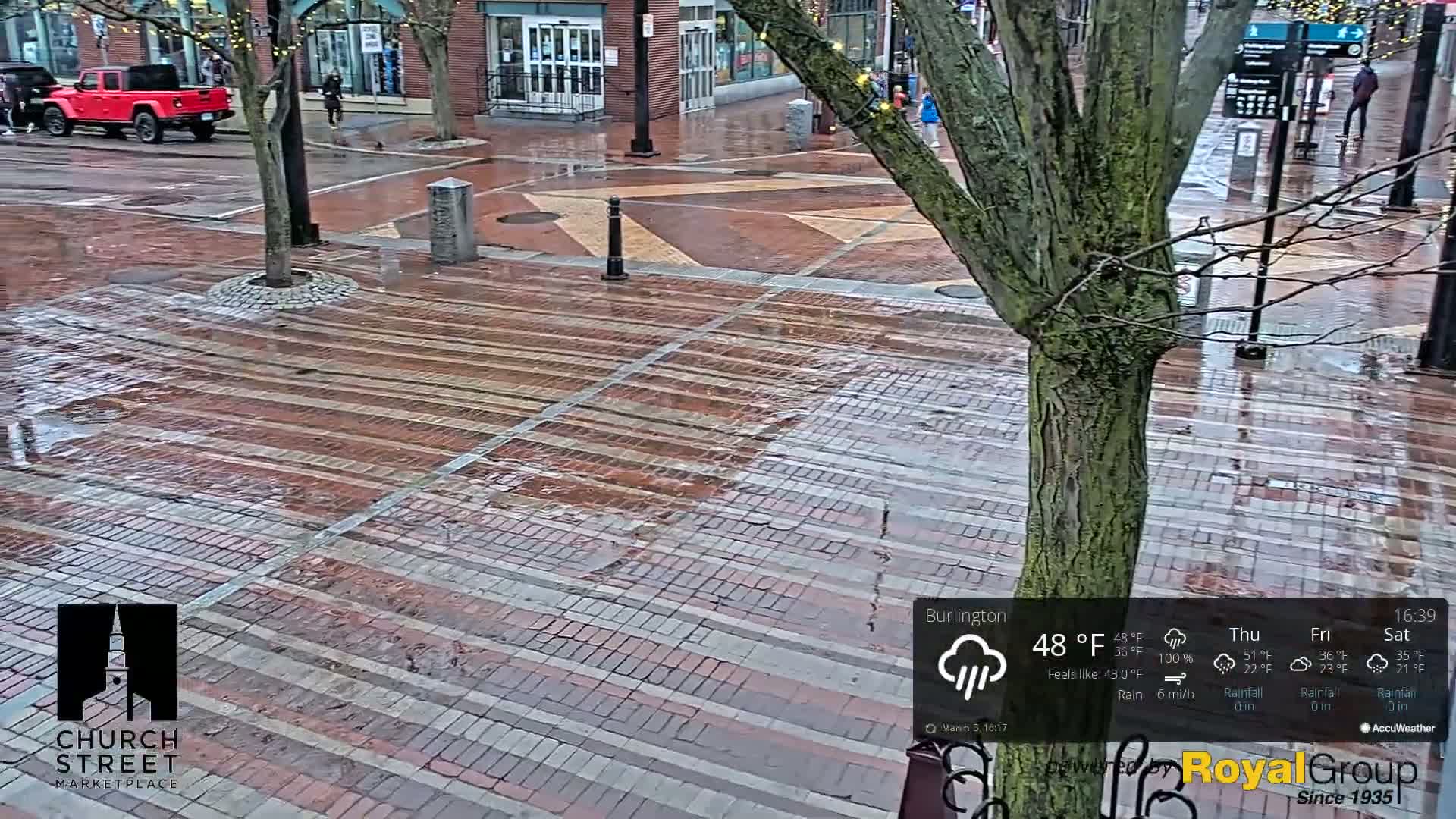 A wet brick pedestrian area is shown with a tree in the center, a red pickup truck in the background, and light rain falling.