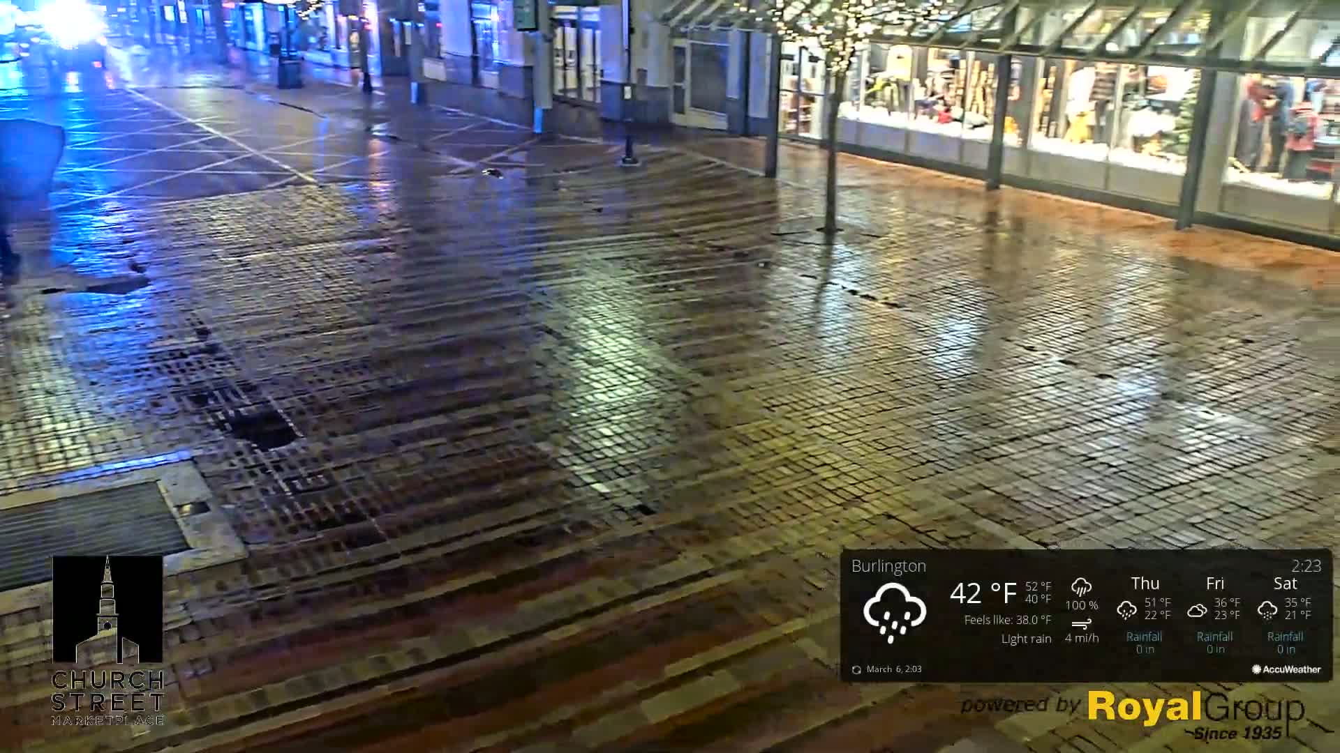 A wet brick-paved street at night is lightly illuminated by streetlights and store windows, with a light rain falling.