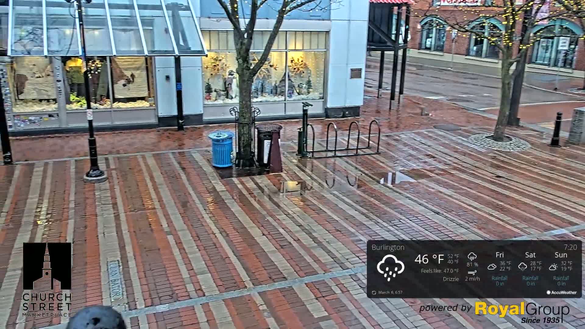 A wet brick paved street scene shows a tree, bike rack, and trash cans under an overcast sky.