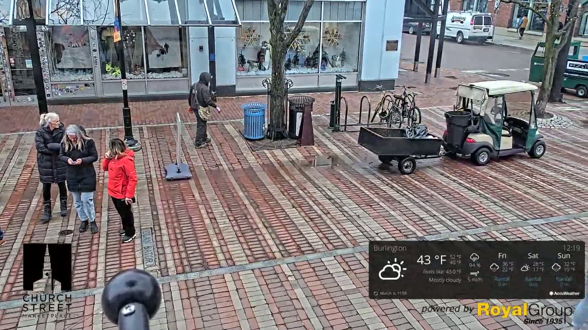 A mostly cloudy day with a temperature of 43°F, showing several people on a brick-paved street, a golf cart with a small trailer, and bicycles parked near a building.