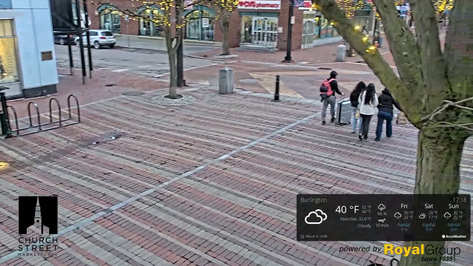 A brick-paved plaza, under cloudy skies, shows a group of people walking near a tree, and some parked cars are visible in the background.
