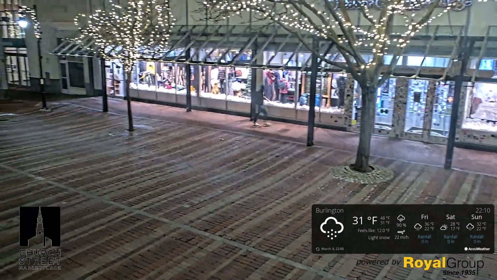 A person walks past a store with holiday lights on a brick paved area during a light snowfall at night.