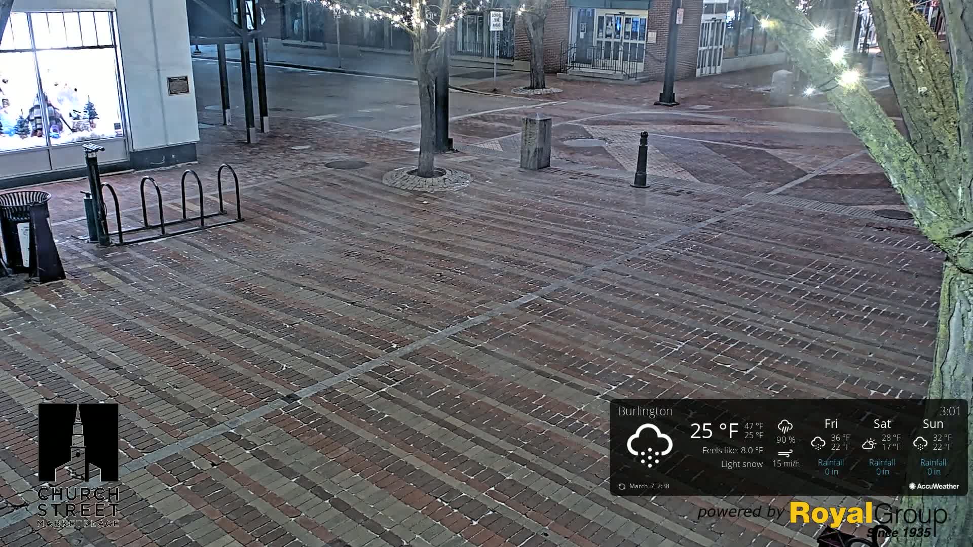 A brick paved town square at night during a light snow with a bike rack and tree strung with lights nearby.