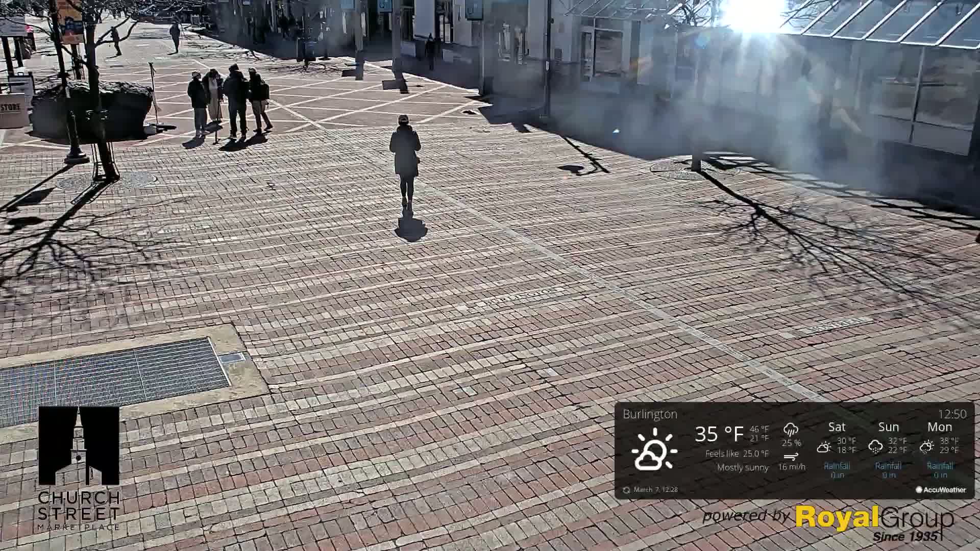 A mostly sunny brick plaza with several people walking and standing in groups, under a bright sun.