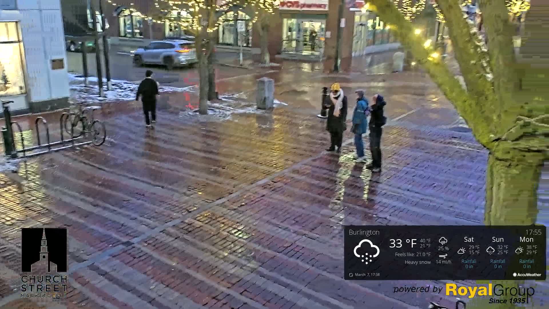 A wet brick plaza with several pedestrians and a bicycle rack, under a heavy snow with a temperature of 33°F.