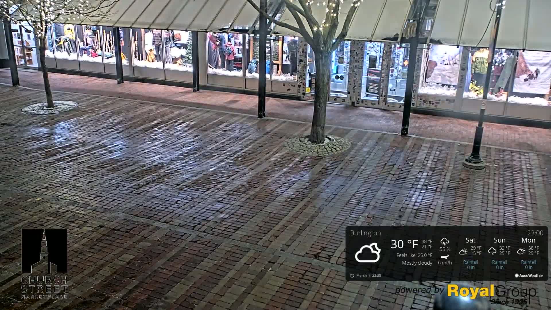 A brick-paved area at night, mostly cloudy, with store fronts displaying winter-themed window displays and a tree with lights.