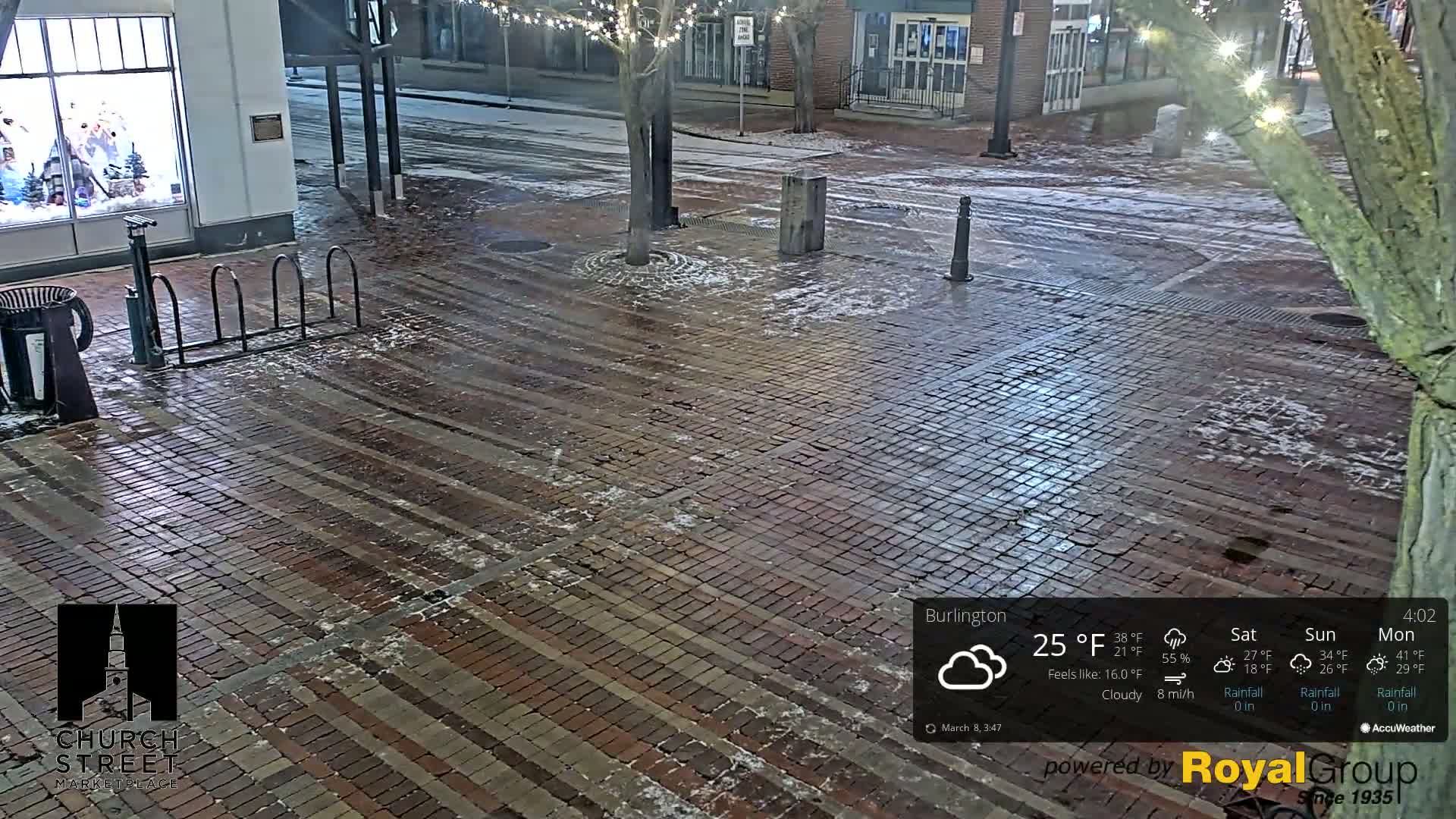 A brick plaza with a bike rack is lightly dusted with snow at night.