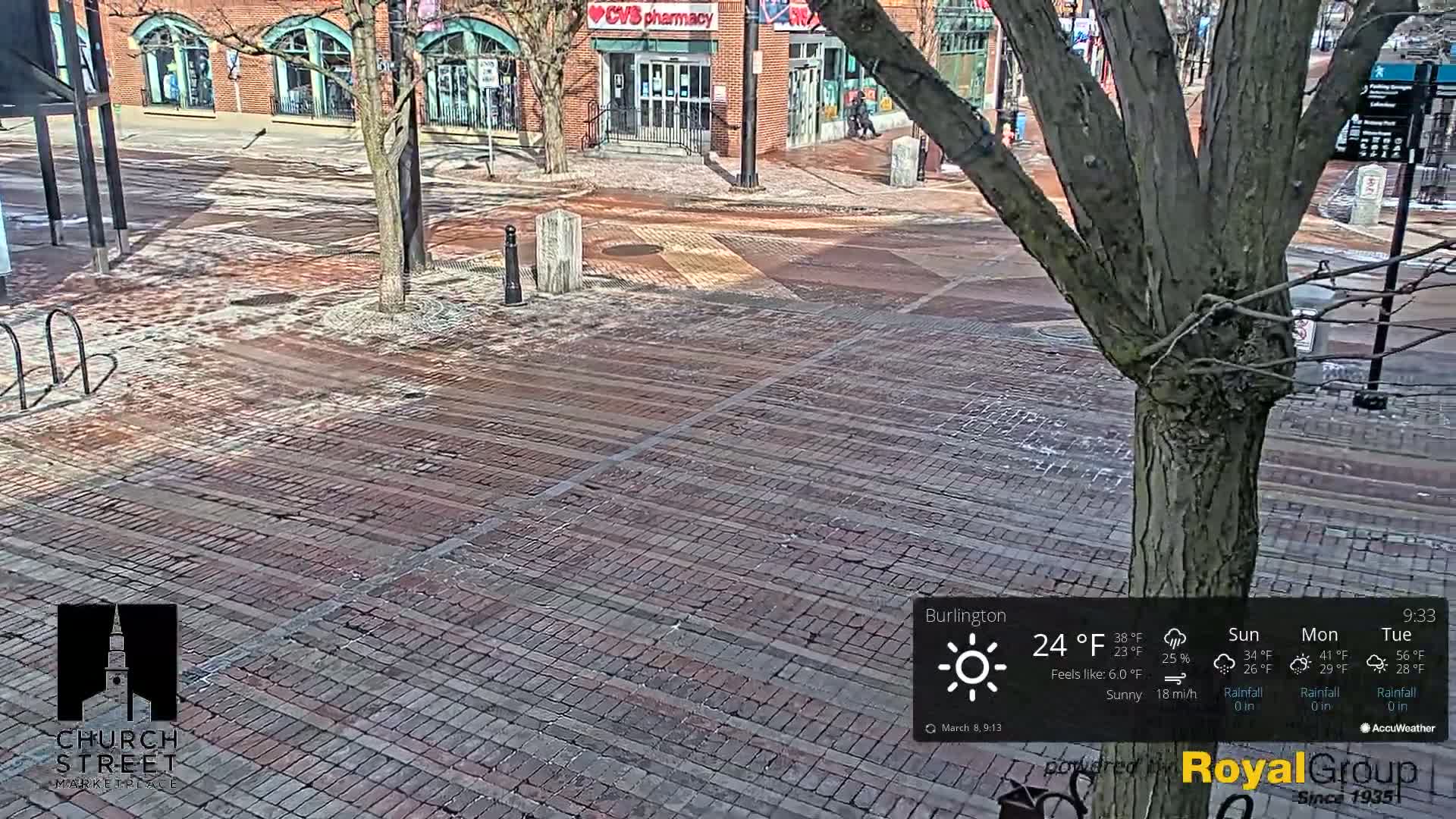 A brick-paved town square, partially covered in snow, is sunny with a light breeze.