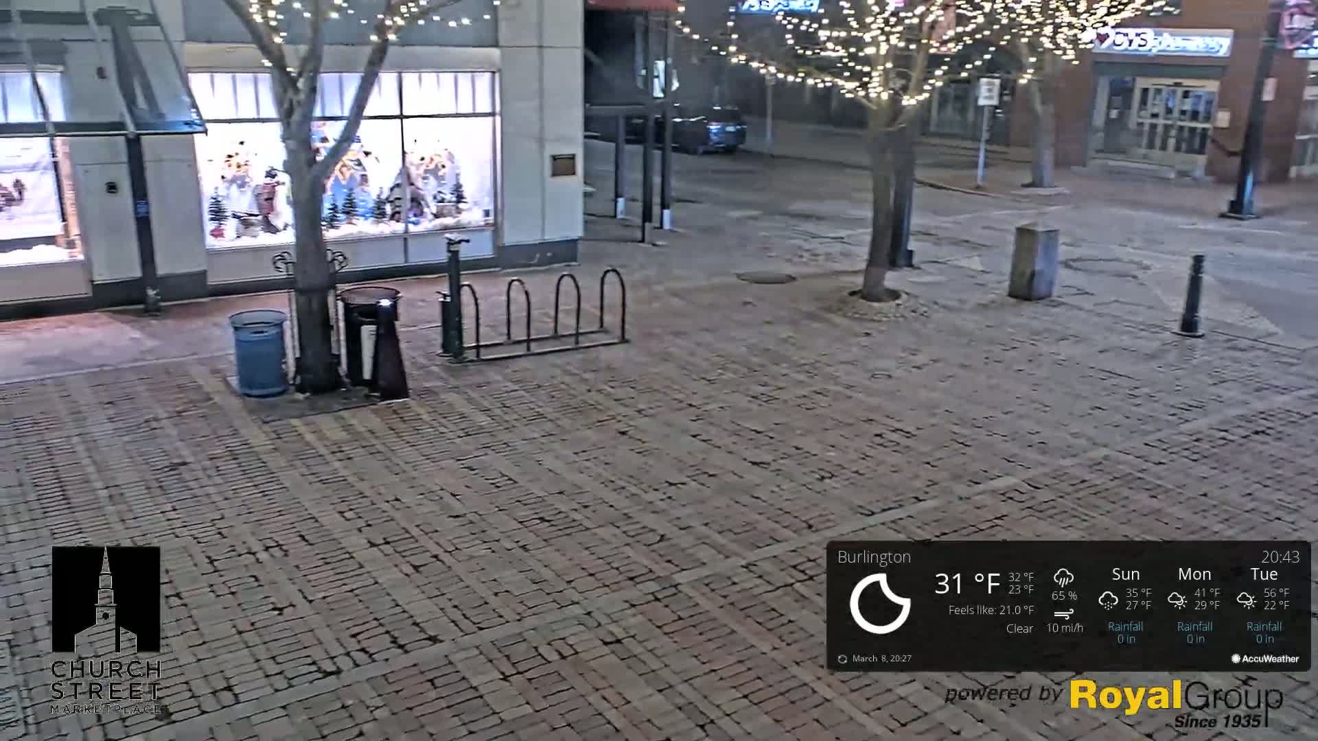 A brick paved area at night, illuminated by streetlights and building lights, shows a bike rack, trash cans, and a water fountain near a store window display with Christmas decorations; the weather is clear with a temperature of 31°F and a wind speed of 10 mi/h.
