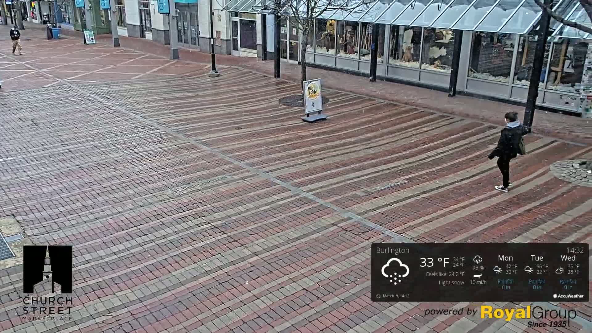 A person walks across a brick-paved street during a light snowfall.