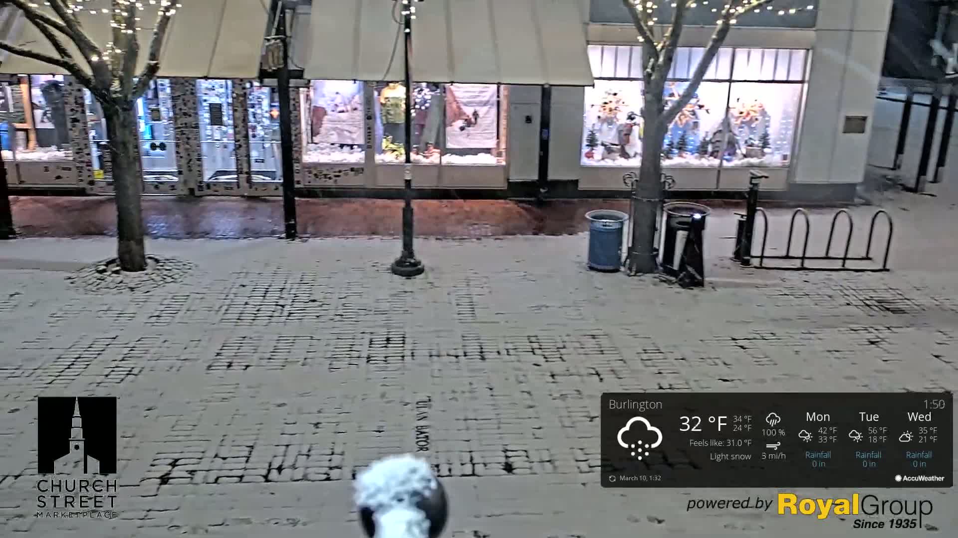 A snow-covered brick street in front of shops with holiday window displays during a light snow.