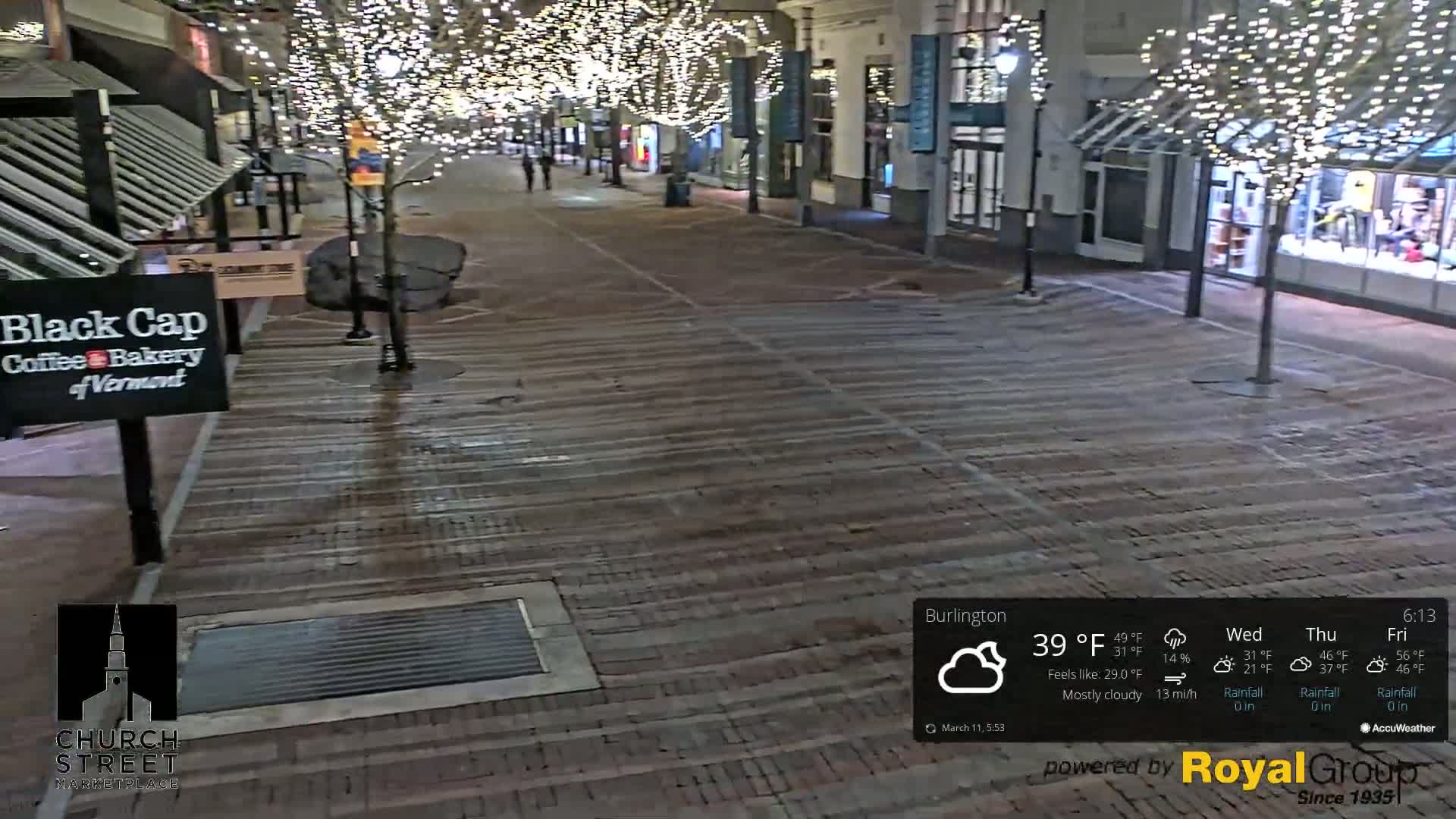 A mostly cloudy night scene shows a brick pedestrian walkway lined with shops and trees decorated with string lights.