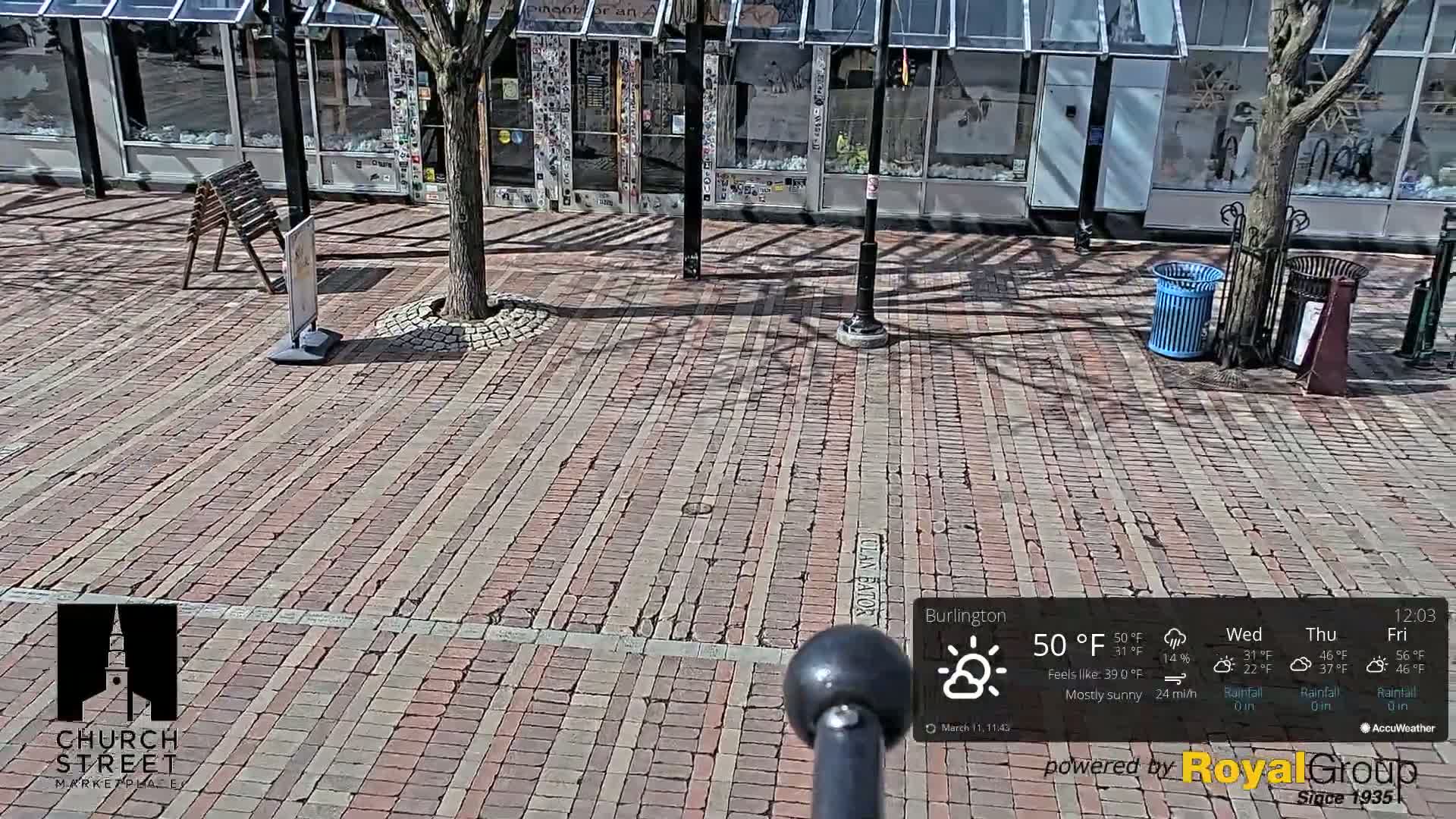 A brick-paved plaza is shown on a mostly sunny day, with a tree and some street furniture visible.