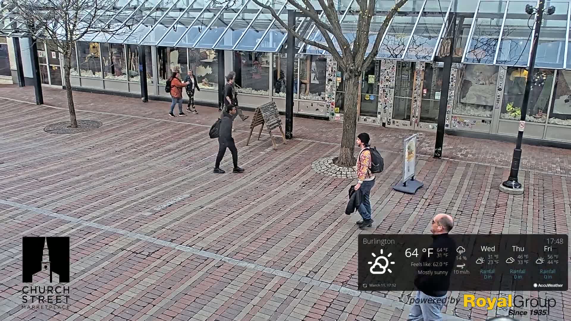 Several people walk on a brick-paved plaza in front of storefronts under a mostly sunny sky.