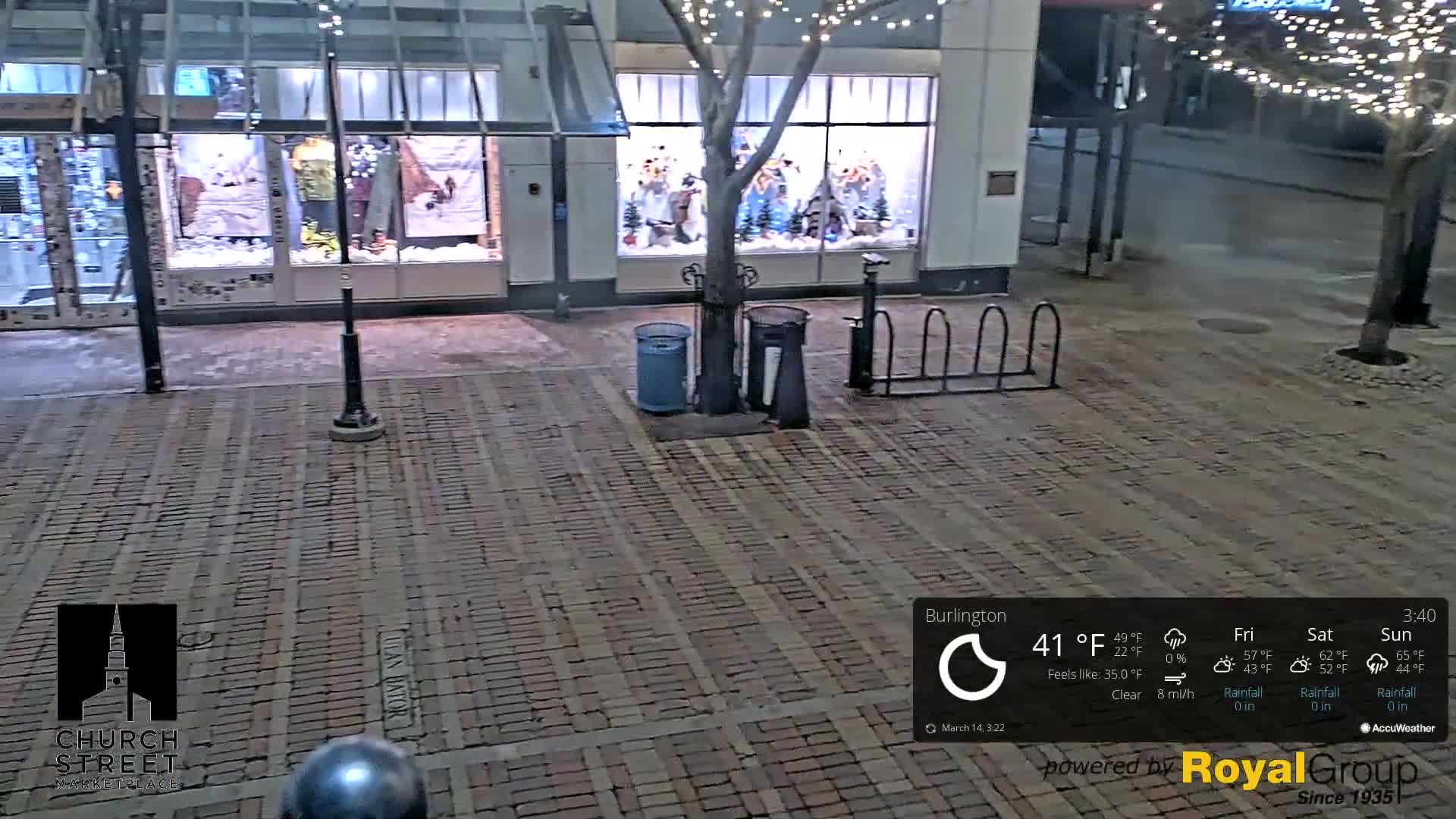 A brick-paved street at night, under clear skies, with a store front displaying holiday decorations and a few trash cans nearby.