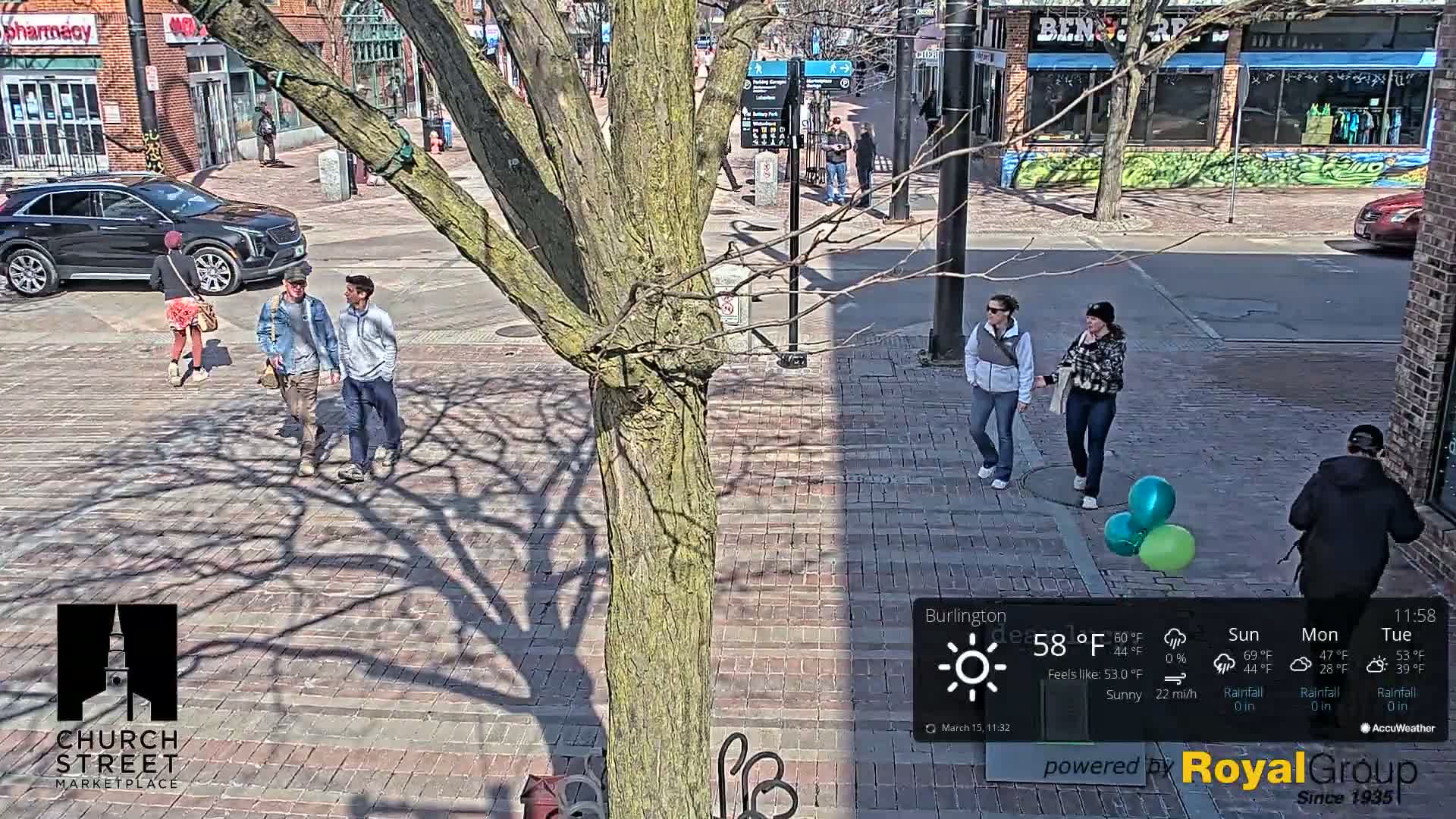 A sunny street scene shows several people walking on a brick pavement, with a large tree casting shadows, and two teal and one green balloon on the ground.