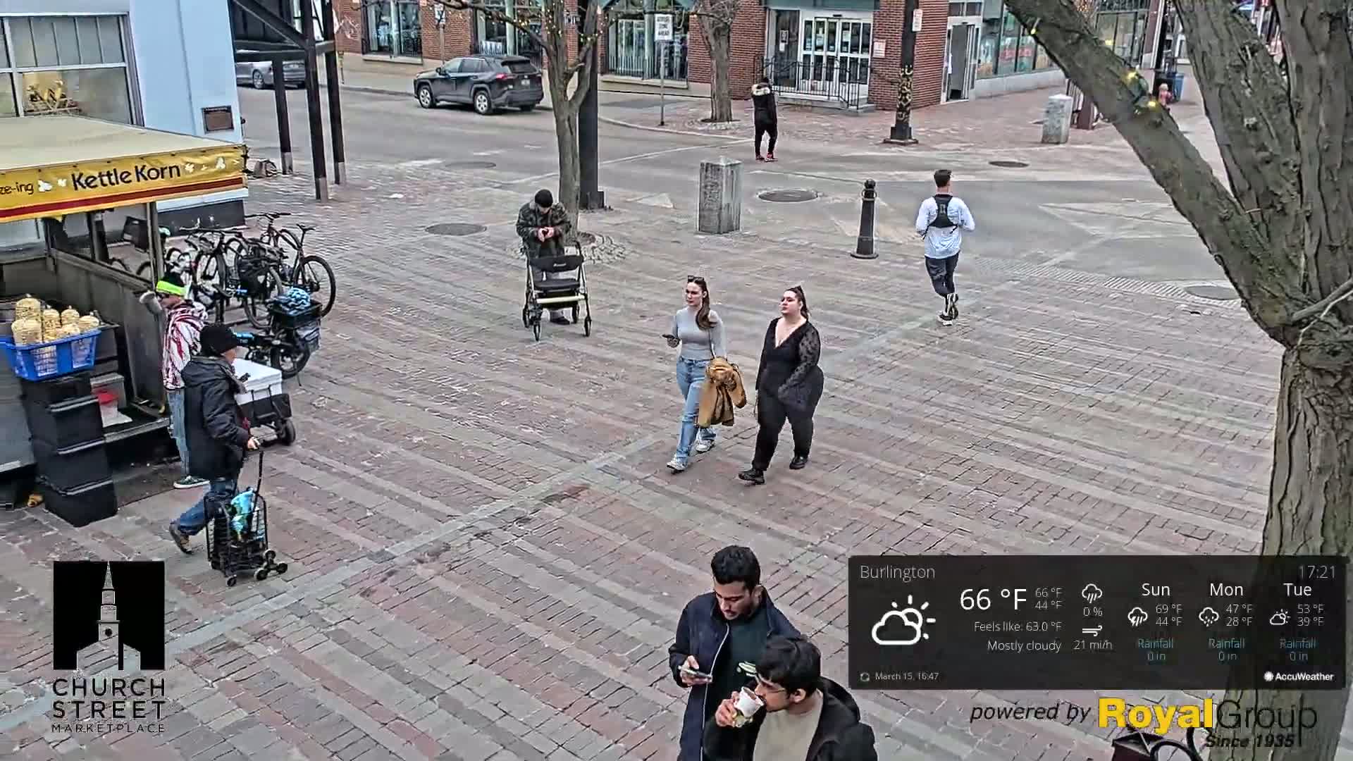 A mostly cloudy, 66°F day shows pedestrians and bicyclists walking and riding on a brick paved street next to a kettle corn stand.