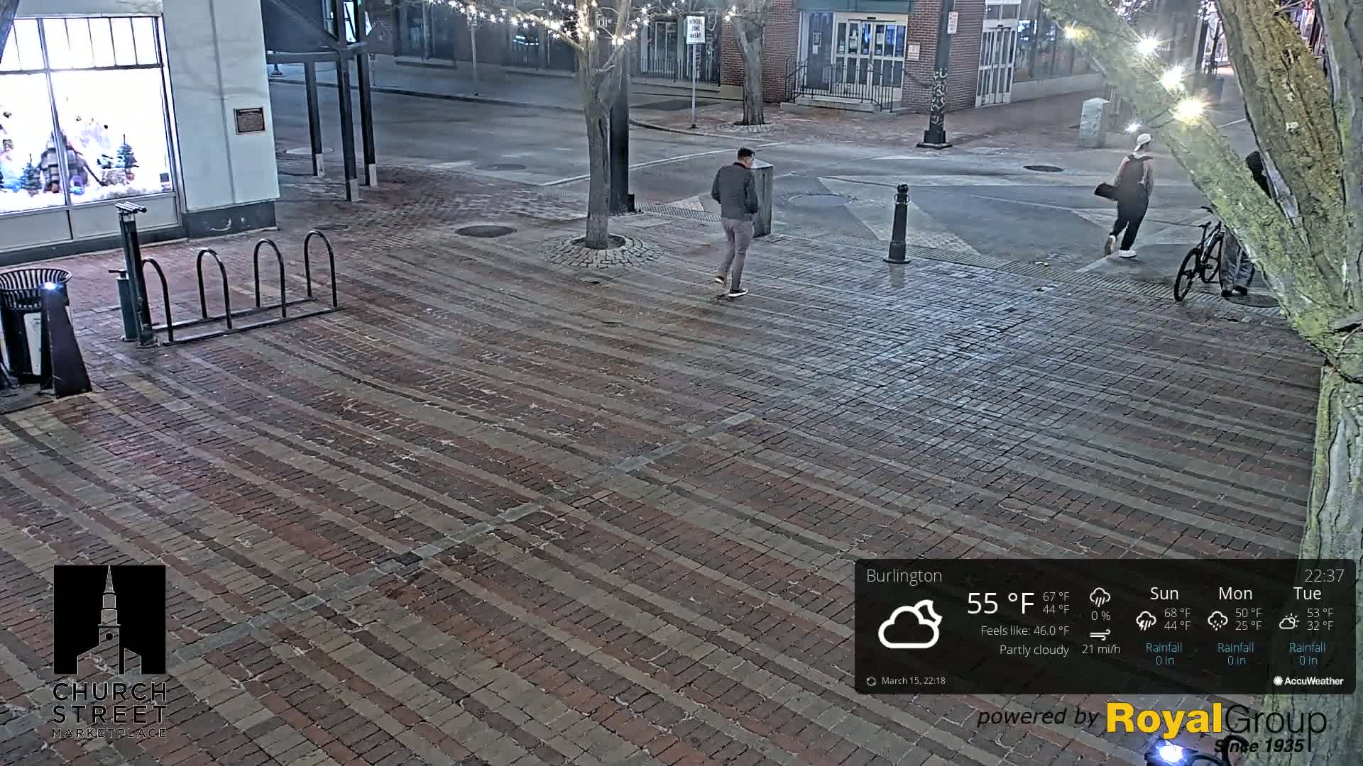 A brick-paved street at night under partly cloudy skies, with two pedestrians and a bicyclist visible.