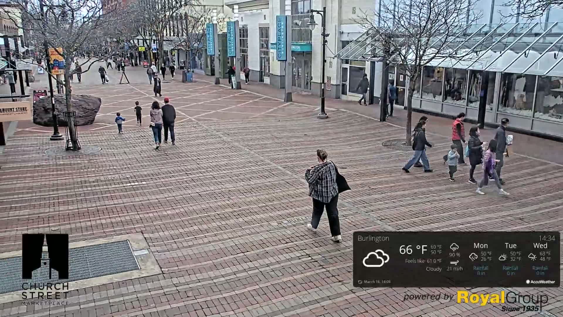 A brick pedestrian walkway in a city is populated with several people walking on a cloudy day.