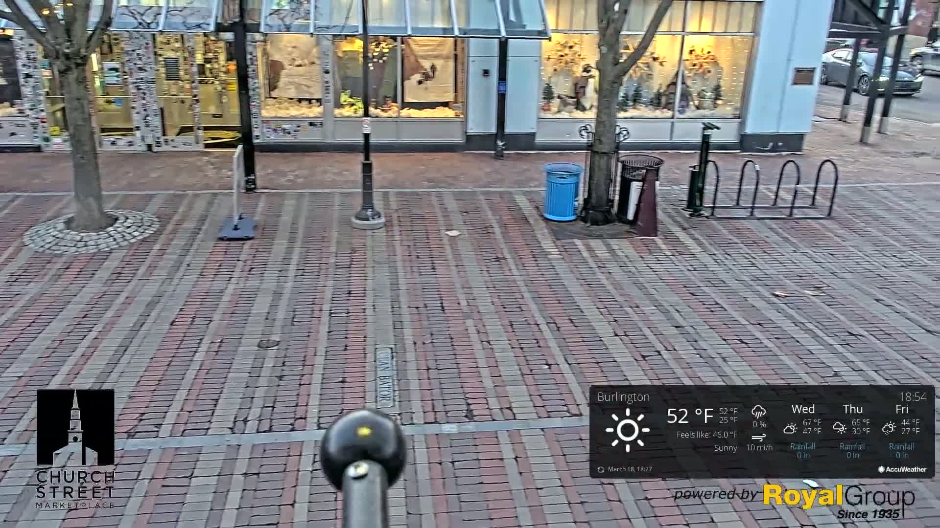 A brick-paved street in front of storefronts on a sunny day with a light wind.