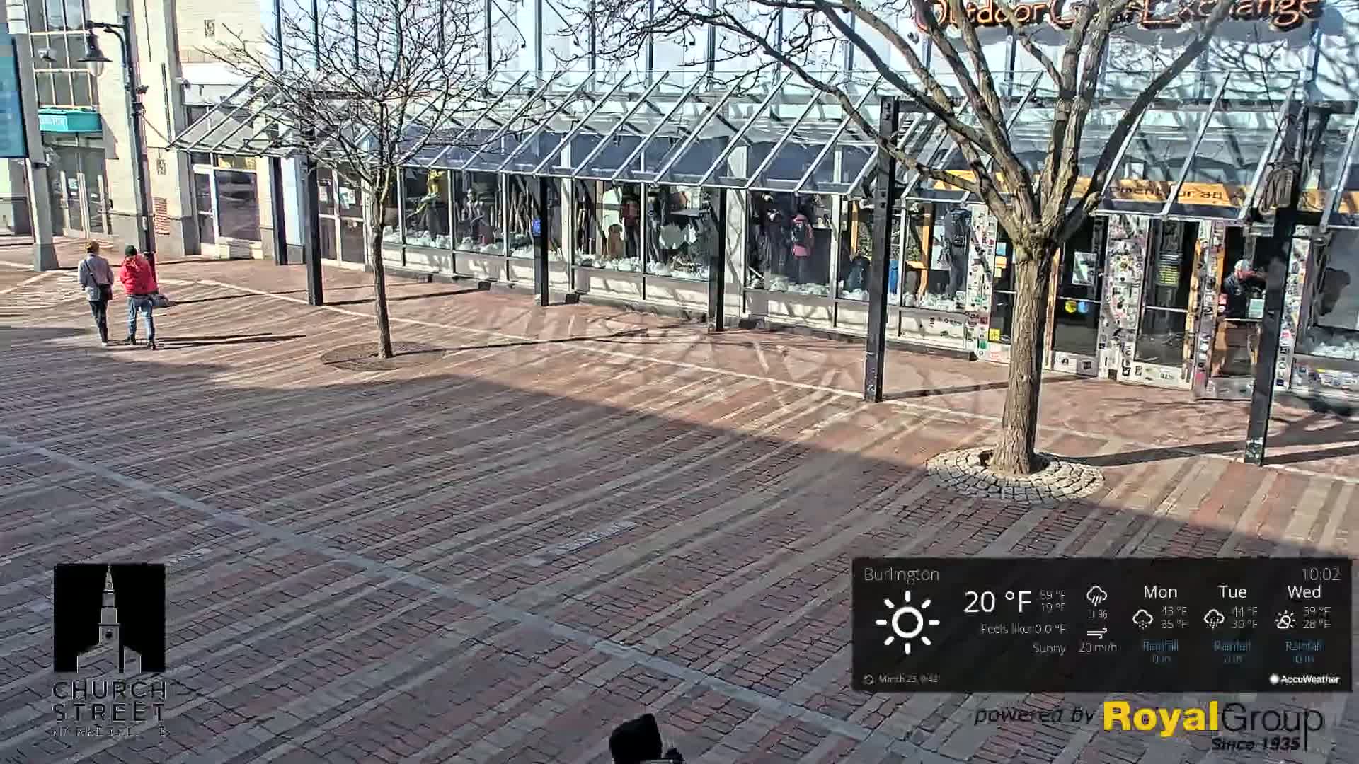 Two people walk on a brick paved street in front of storefronts under a sunny sky.