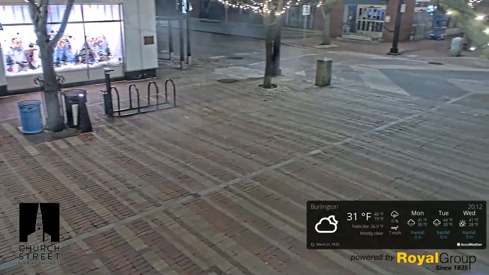 A mostly clear night scene shows a brick paved area with bike racks, trash cans, and a tree near a storefront.